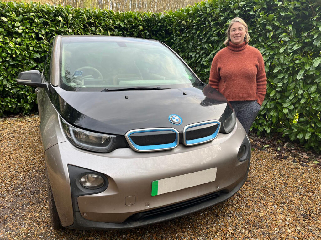 Vicky Edmonds, wearing a rust-coloured sweater and grey trousers, standing next to her silver and black BMW i3